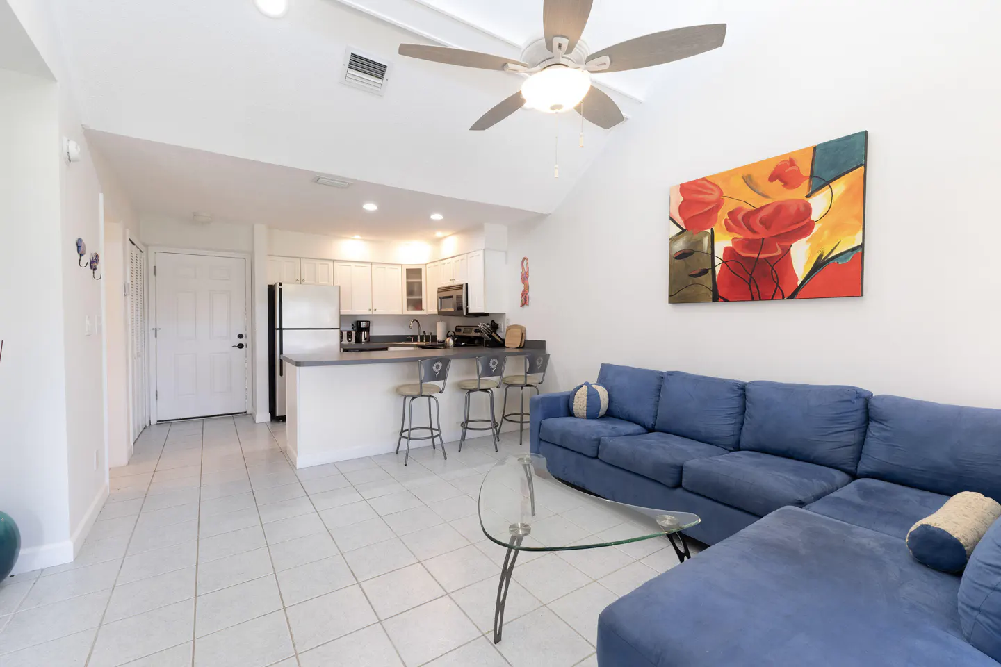 Bright living space with white tile floors, a blue sectional sofa, and an open kitchen with white cabinets. A ceiling fan hangs above.