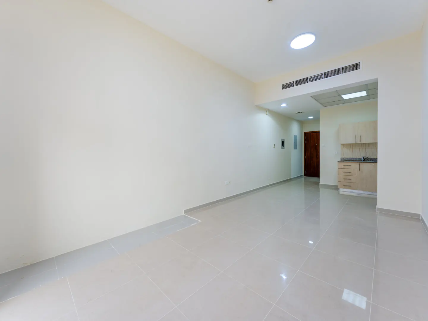 Empty room with beige walls, glossy tiled floors, and a small kitchenette with light wood cabinets in the background.