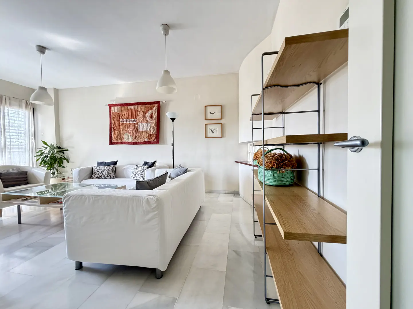 Bright living room with white sofa, marble floors, and a wooden shelf with a green basket. A tapestry hangs on the wall.