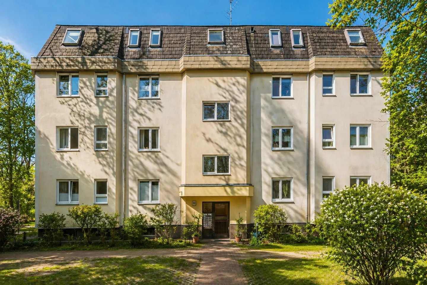 Exterior of a three-story apartment building with a brown roof and a yellow facade, surrounded by green trees and bushes.