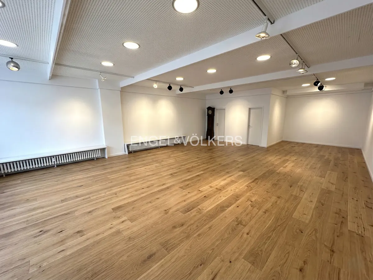 Bright, empty room with light wood floors, white walls, and a white perforated ceiling with recessed lighting. Radiators line the walls. A grandfather clock stands in the back.