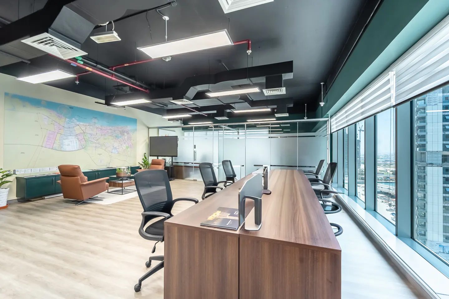 Bright office space with a long wooden desk, black chairs, and large windows overlooking city buildings. A map and lounge area are in the background.