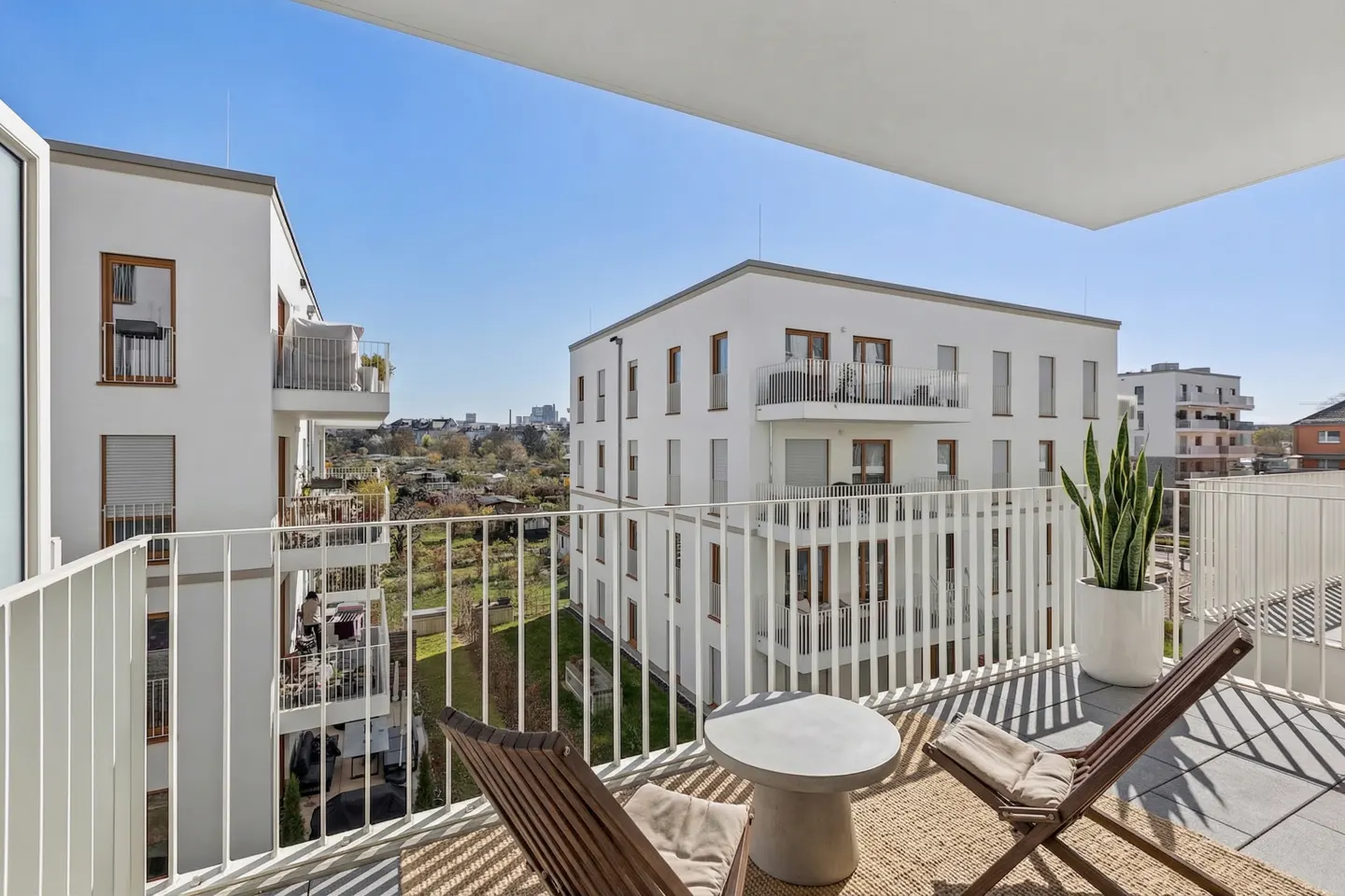 Balcony view with two wooden chairs, a small table, and a potted plant. White buildings and a blue sky are in the background.