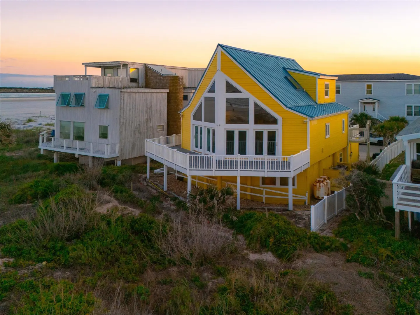 Bright yellow beach house with a blue metal roof and large windows, featuring a wrap-around deck at sunset.