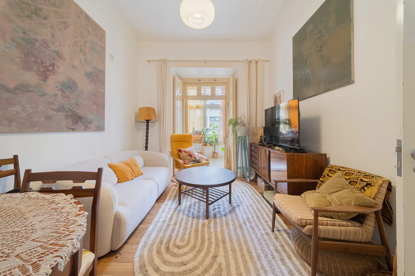 Bright living room with white walls, wood floors, and a patterned rug. A white sofa, wooden chairs, and a TV stand furnish the space. Artwork hangs on the walls.