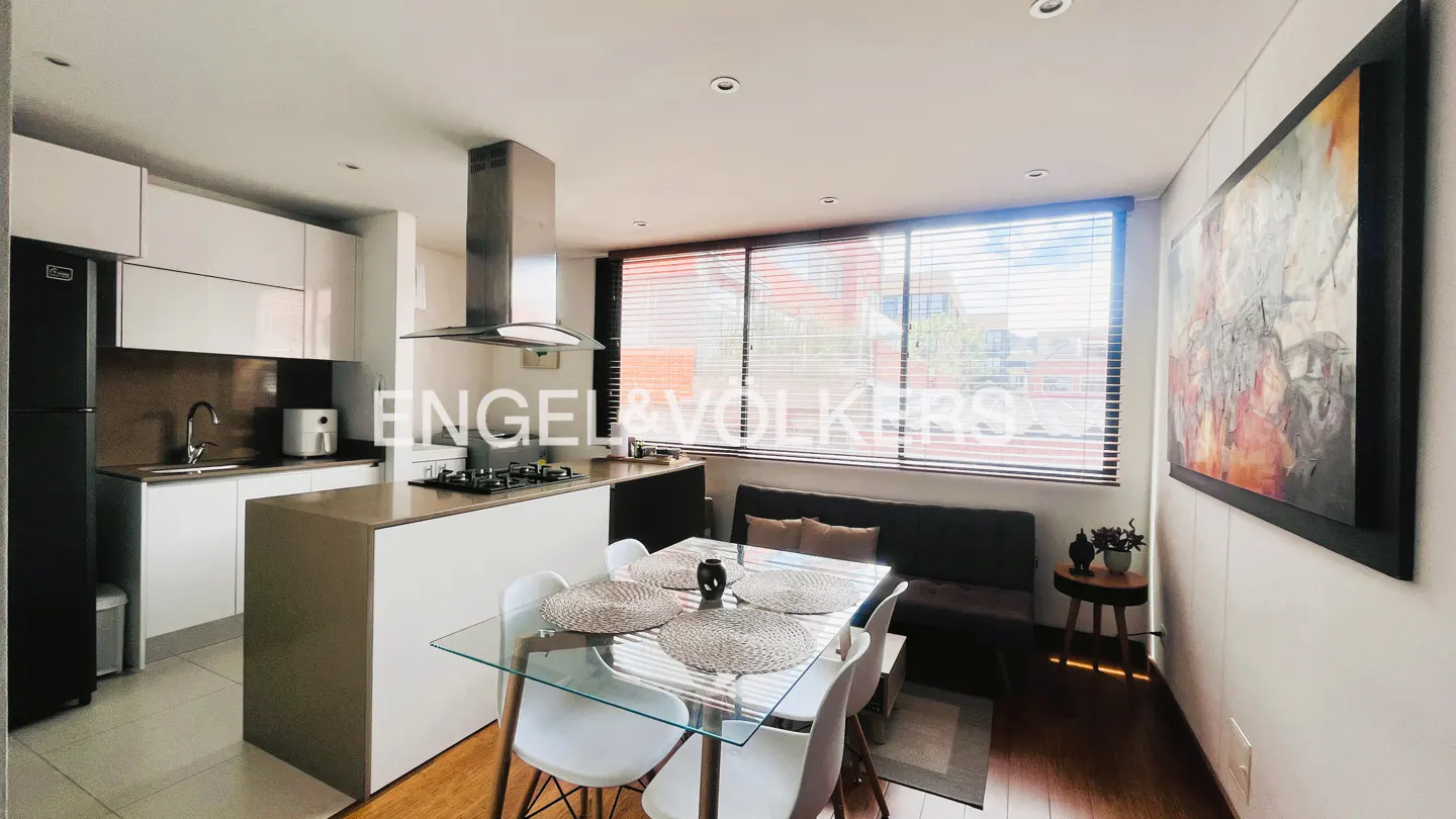 Bright, modern apartment interior with white kitchen, glass table, and dark sofa by window. Abstract art hangs on the wall.