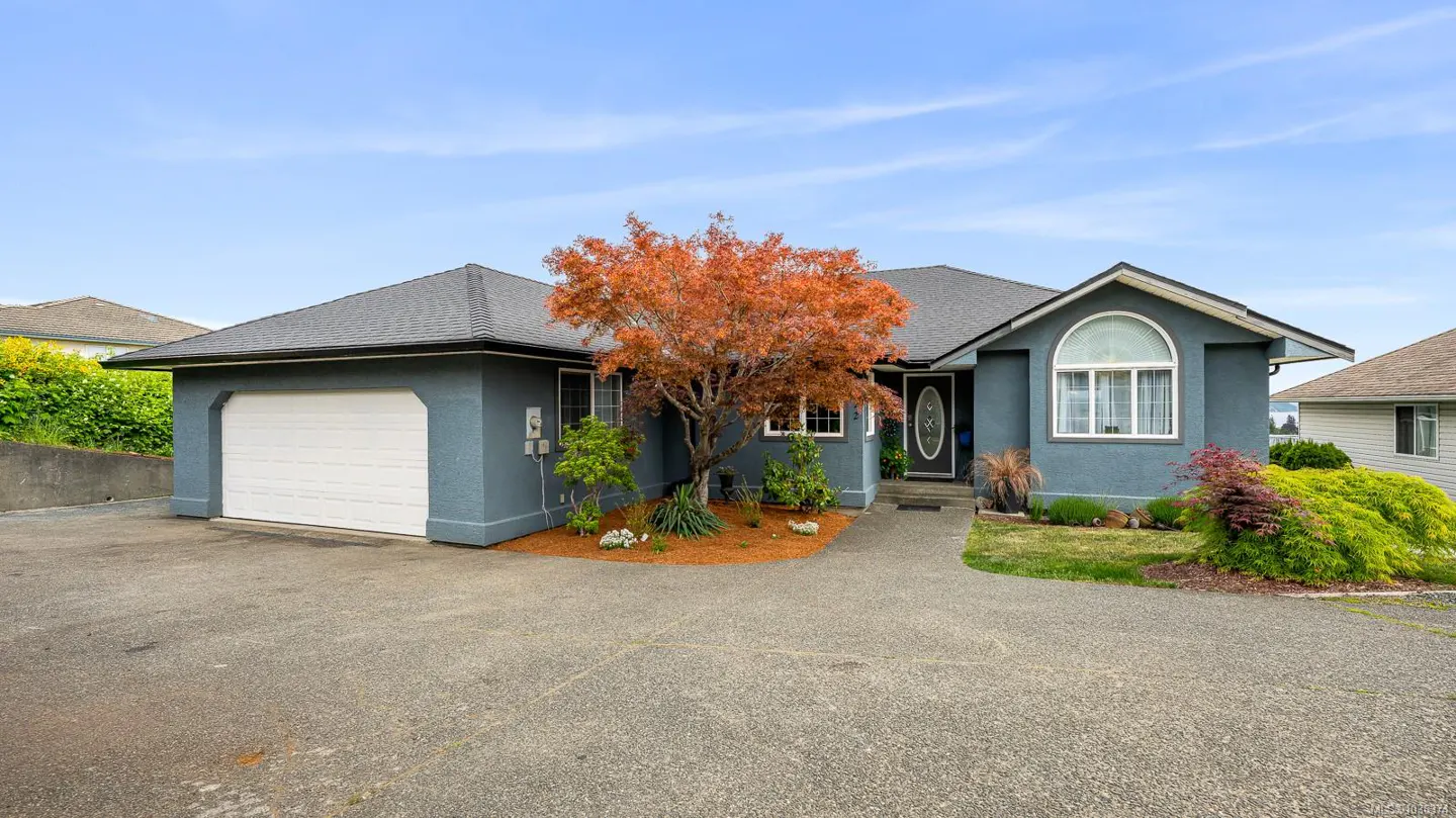 Exterior view of a one-story blue house with a gray roof, white garage door, and a red-leaved tree in the front yard.