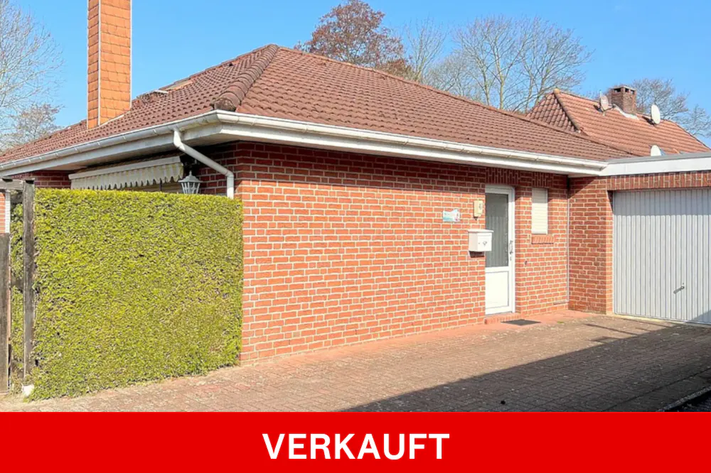 A single-story brick house with a red tile roof and a white garage door. A green hedge is on the left. "VERKAUFT" is written in red at the bottom.