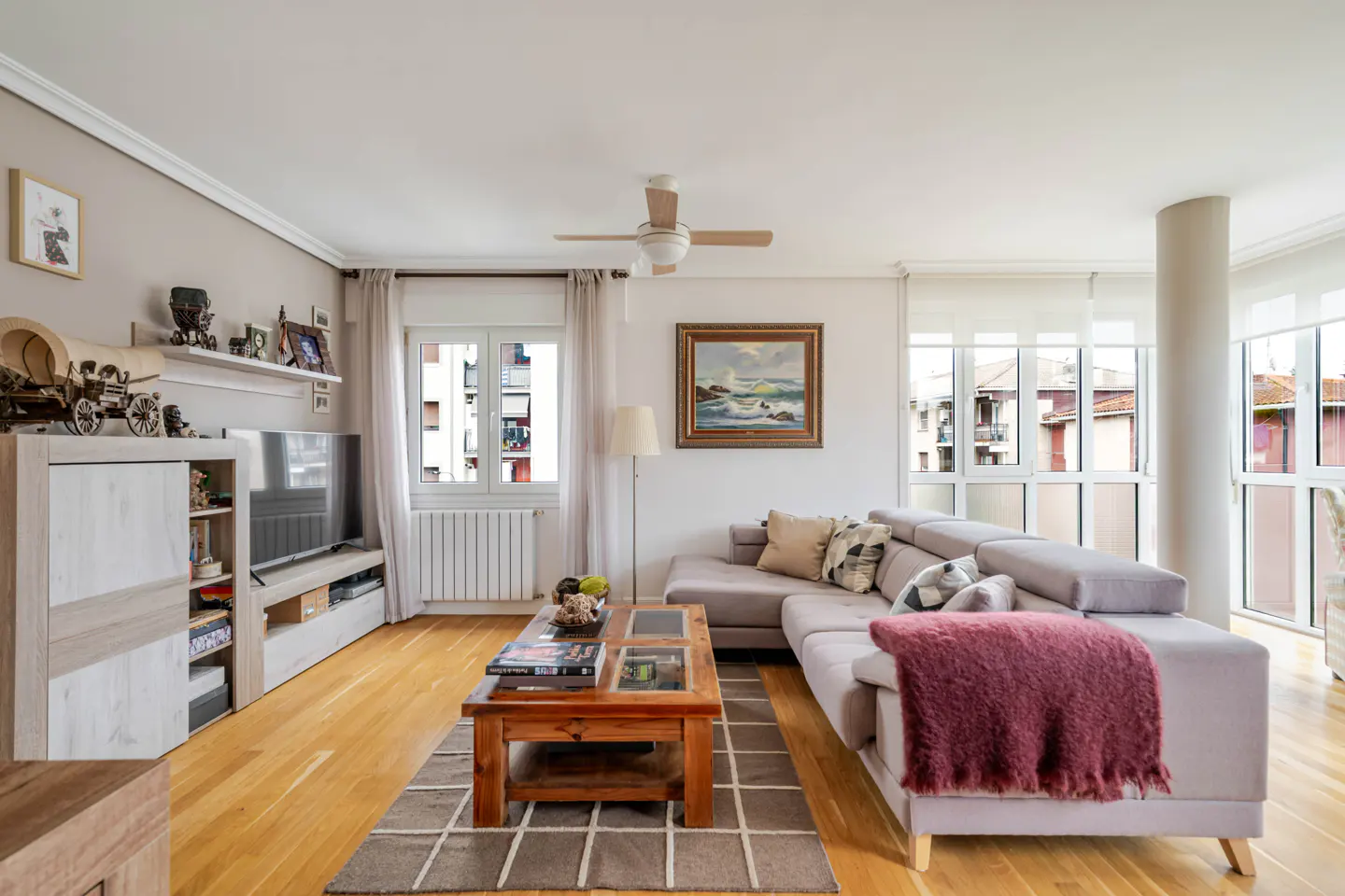 Bright living room with light wood floors, a grey sectional sofa with a maroon throw, and a wooden coffee table.