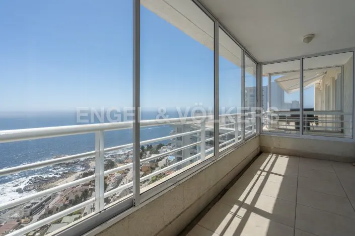 Balcony view of the ocean through glass windows and a white railing. The floor is tiled and light brown.