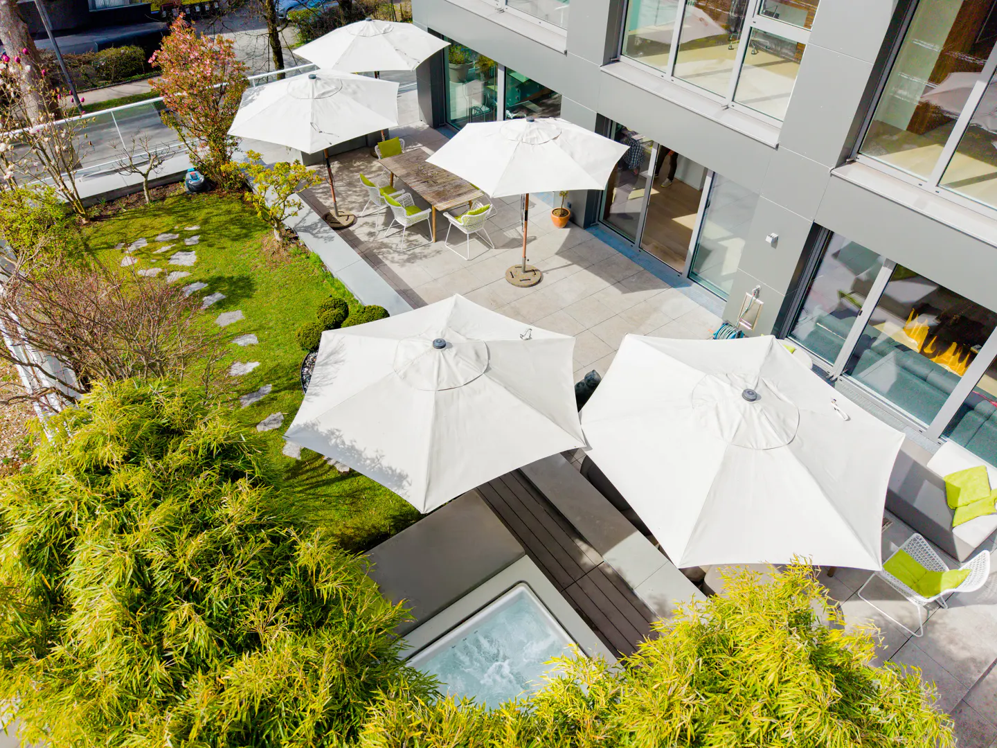 Aerial view of a modern patio with white umbrellas, a hot tub, green foliage, and a dining table with chairs.
