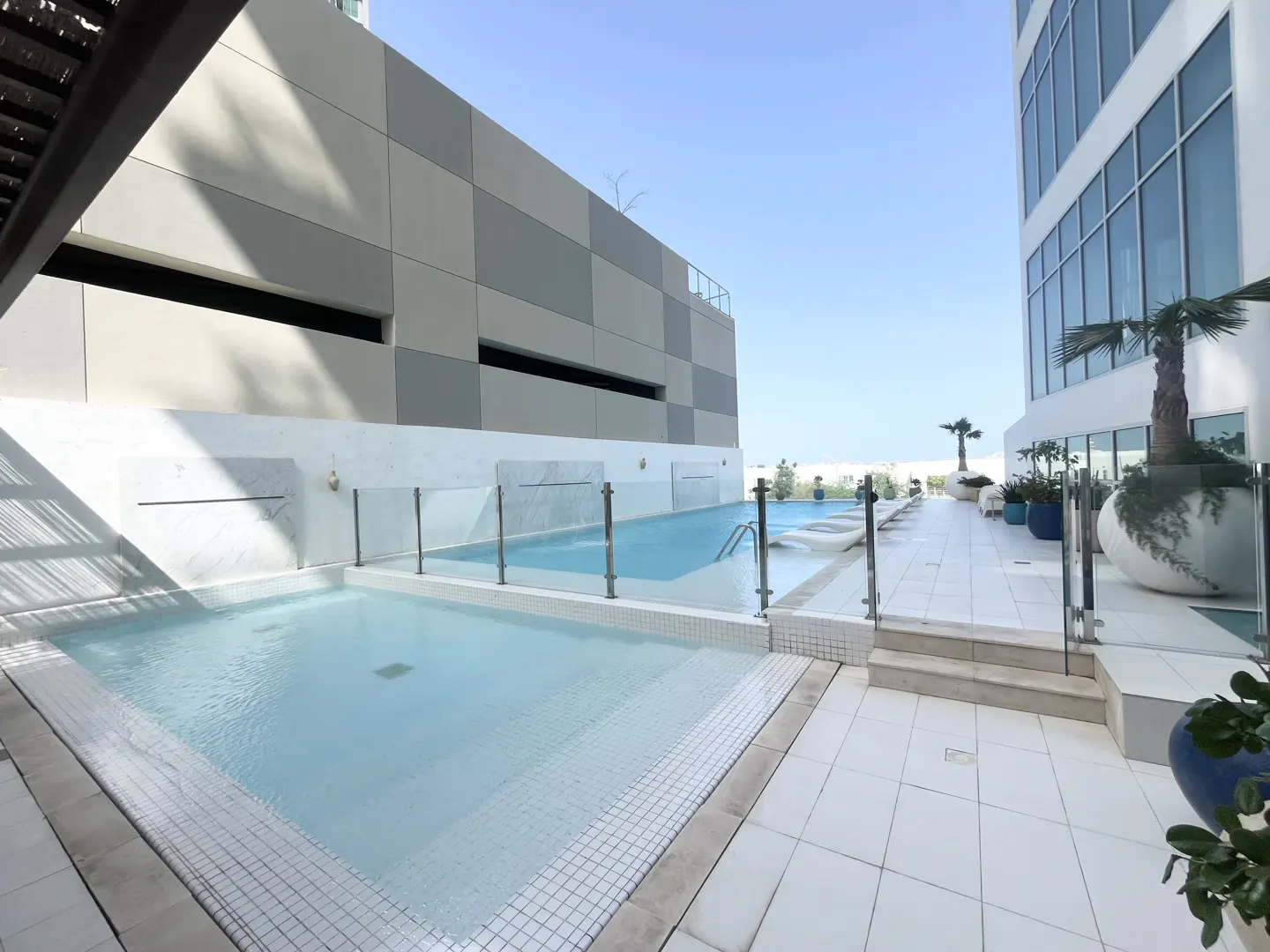 Rooftop pool area with light blue water, white tile, and modern buildings under a clear blue sky.
