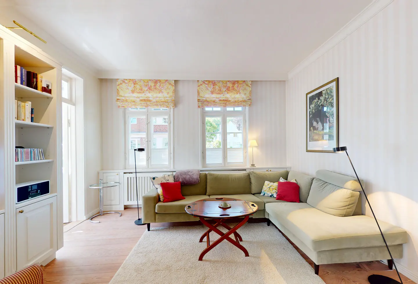 Bright living room with a light green sectional sofa, red accent pillows, a round wooden coffee table, and floral blinds.