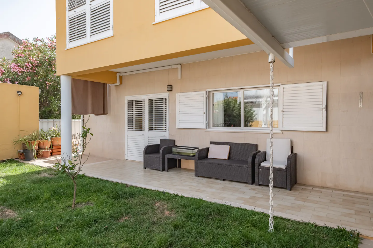Backyard patio with gray outdoor furniture, a small tree, and a yellow two-story house in the background.