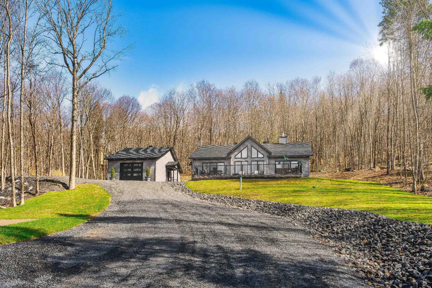 A modern gray house and garage sit on a green lawn, surrounded by bare trees under a bright blue sky. A gravel driveway leads to the buildings.