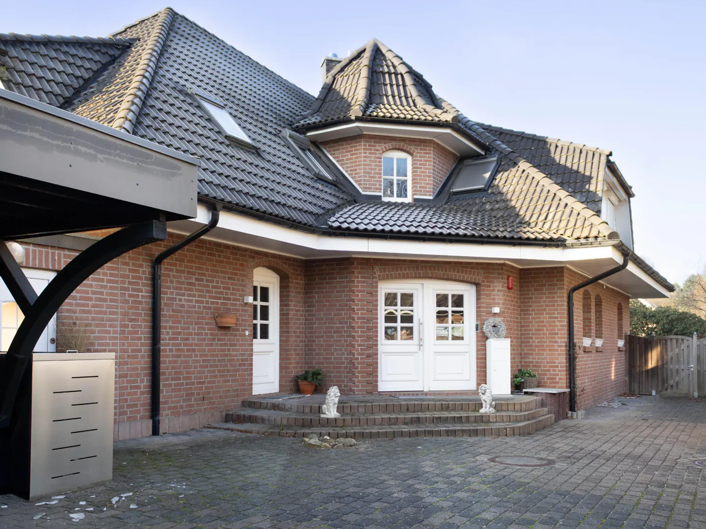 Brick house with a dark gray tiled roof, white doors, and a paved driveway. Lion statues flank the steps to the entrance.