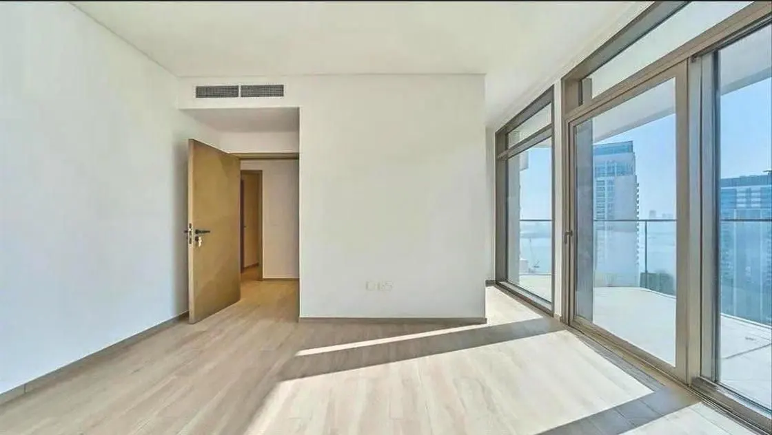 An empty room with light wood floors, white walls, and large windows with a city view. A door is slightly ajar.