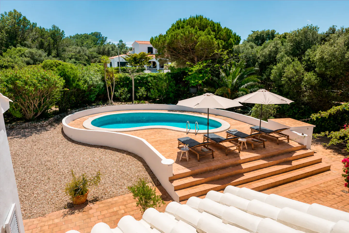 Overhead shot of a kidney-shaped pool with blue water, surrounded by lounge chairs, umbrellas, and lush greenery.