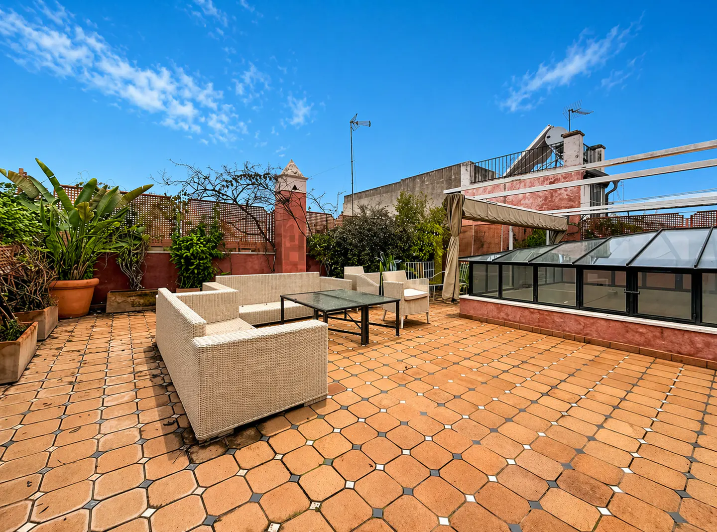 Rooftop patio with wicker furniture, glass-topped table, and tiled floor. Plants line the perimeter, with a blue sky above.