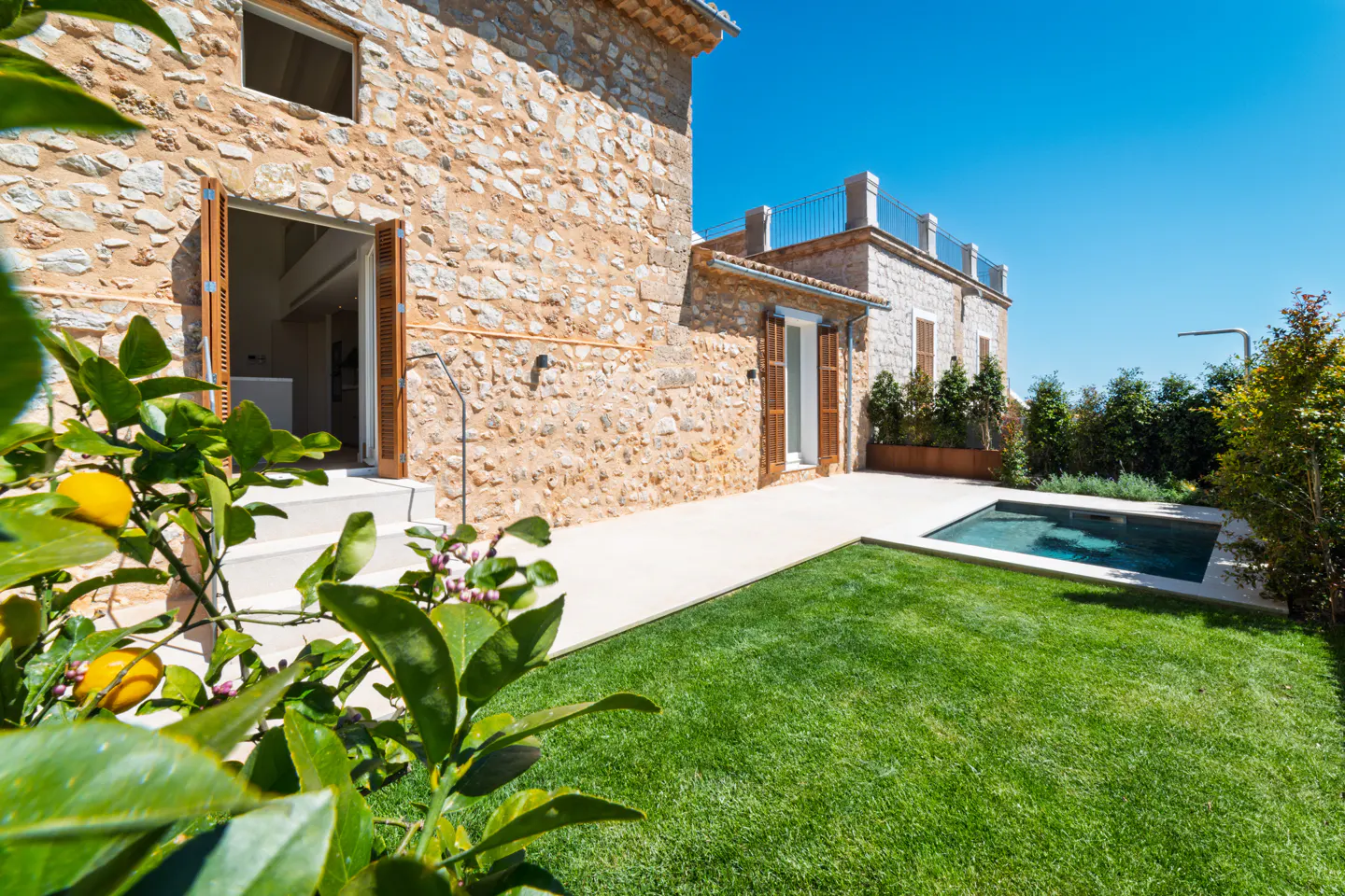 Exterior view of a stone house with a small pool and green lawn under a clear blue sky. Lemon tree in the foreground.