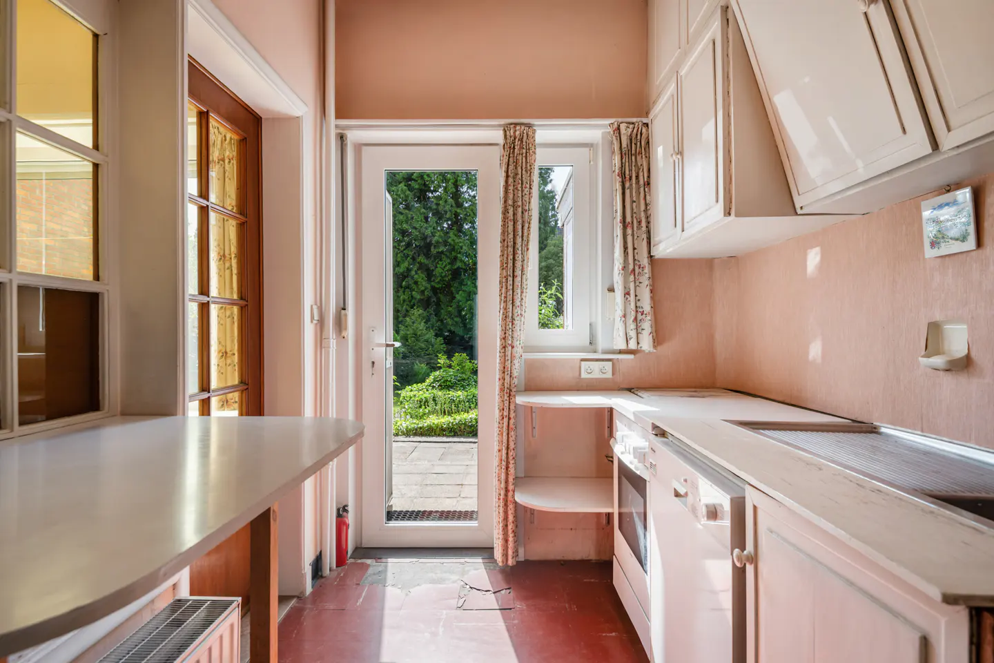 A pink kitchen with white cabinets, a white countertop, and a door leading to a garden.