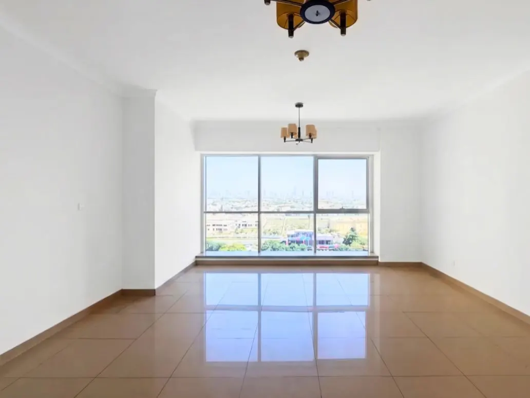 Bright, empty room with white walls, brown tile floor, and large window showing a city view. Two light fixtures on the ceiling.