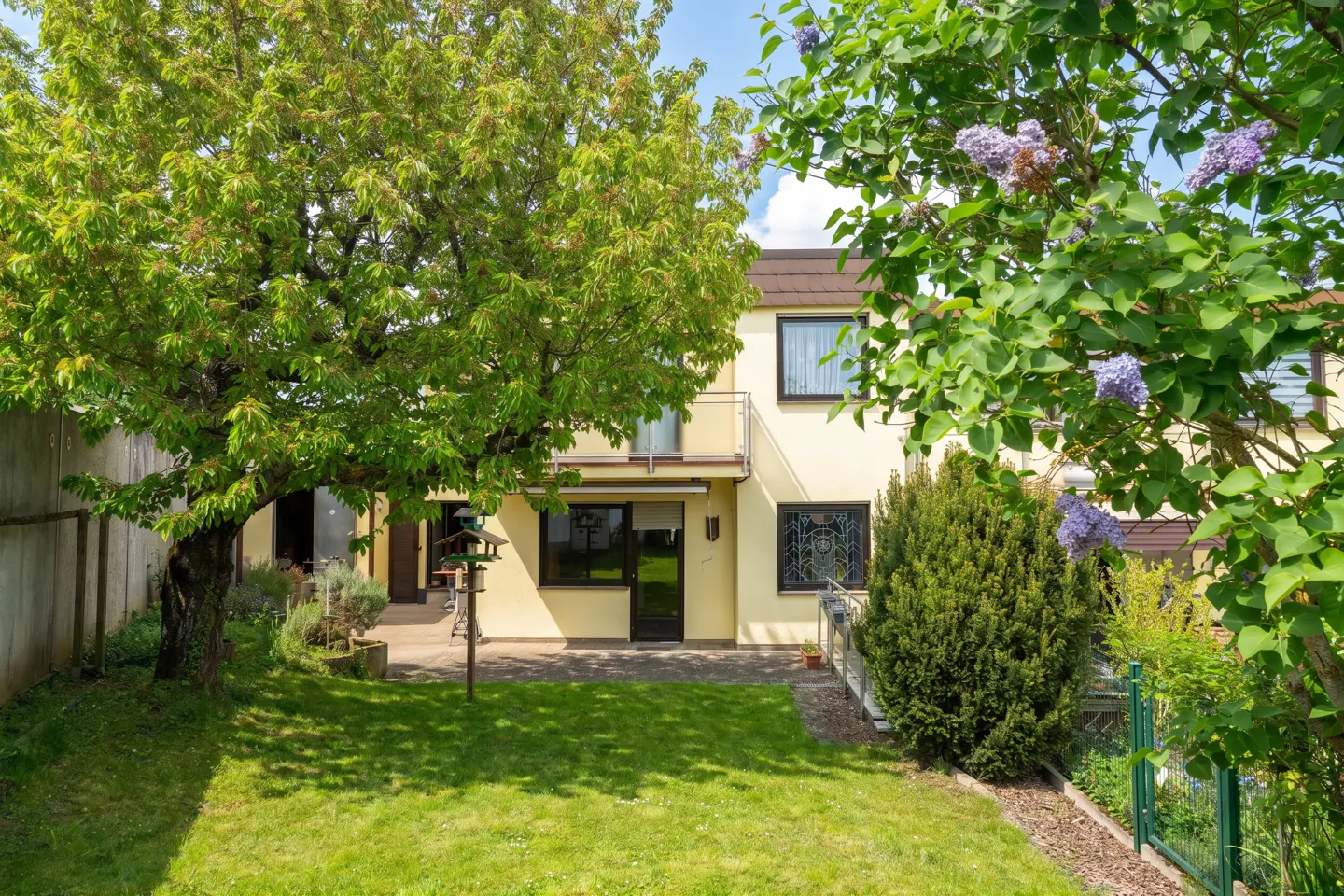 Backyard view of a two-story, light yellow house with a green lawn, trees, and a bird feeder.