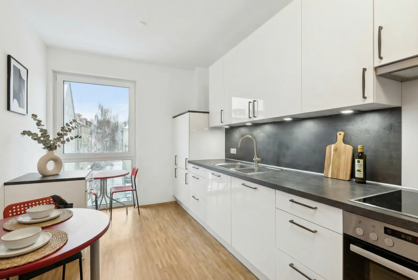 Bright kitchen with white cabinets, dark countertops, and wood floors. A red table and chairs are near a window. A vase with greenery sits on a counter.