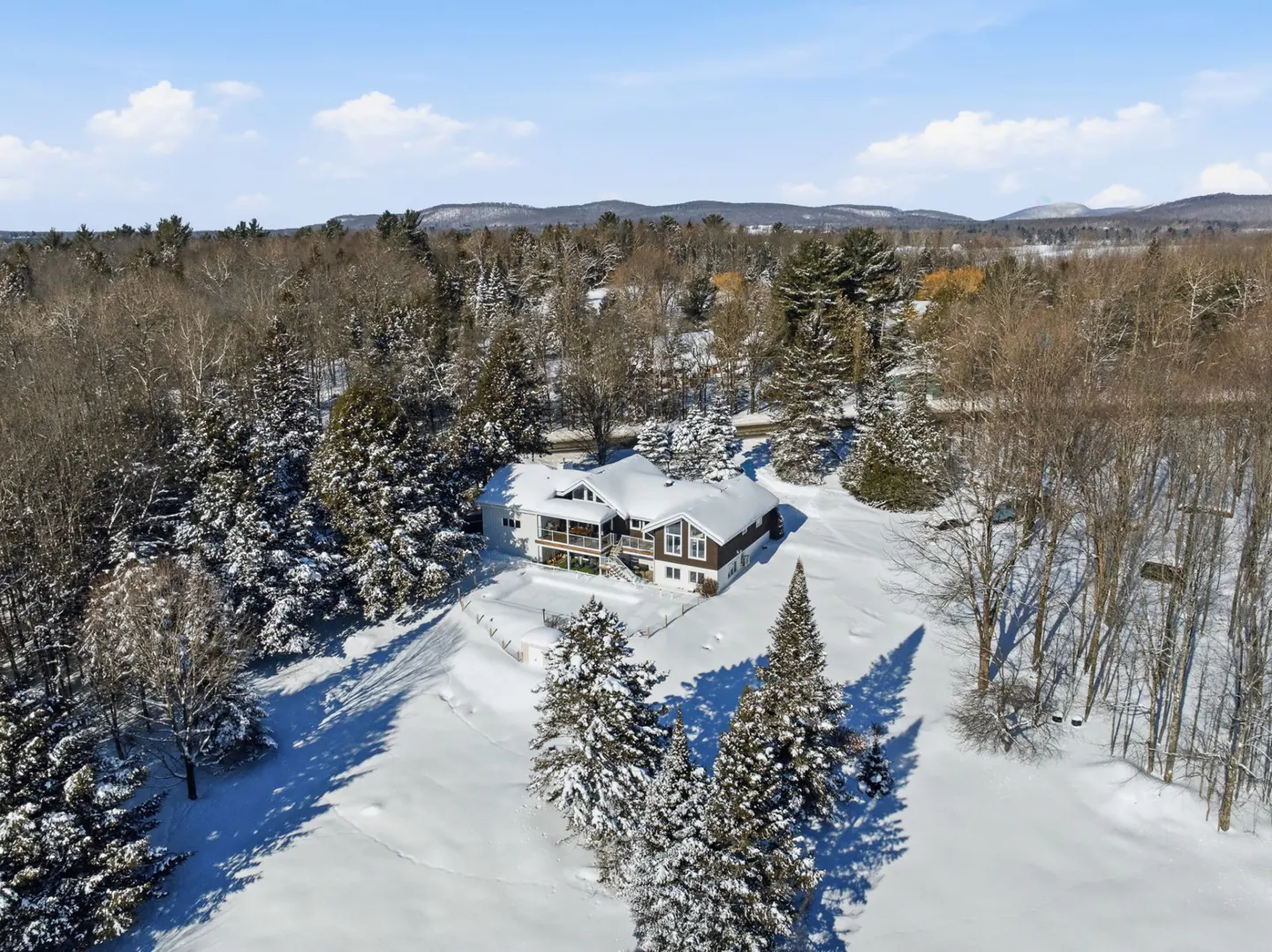 A snow-covered house with a balcony is surrounded by trees and snow. The sky is blue with some clouds.