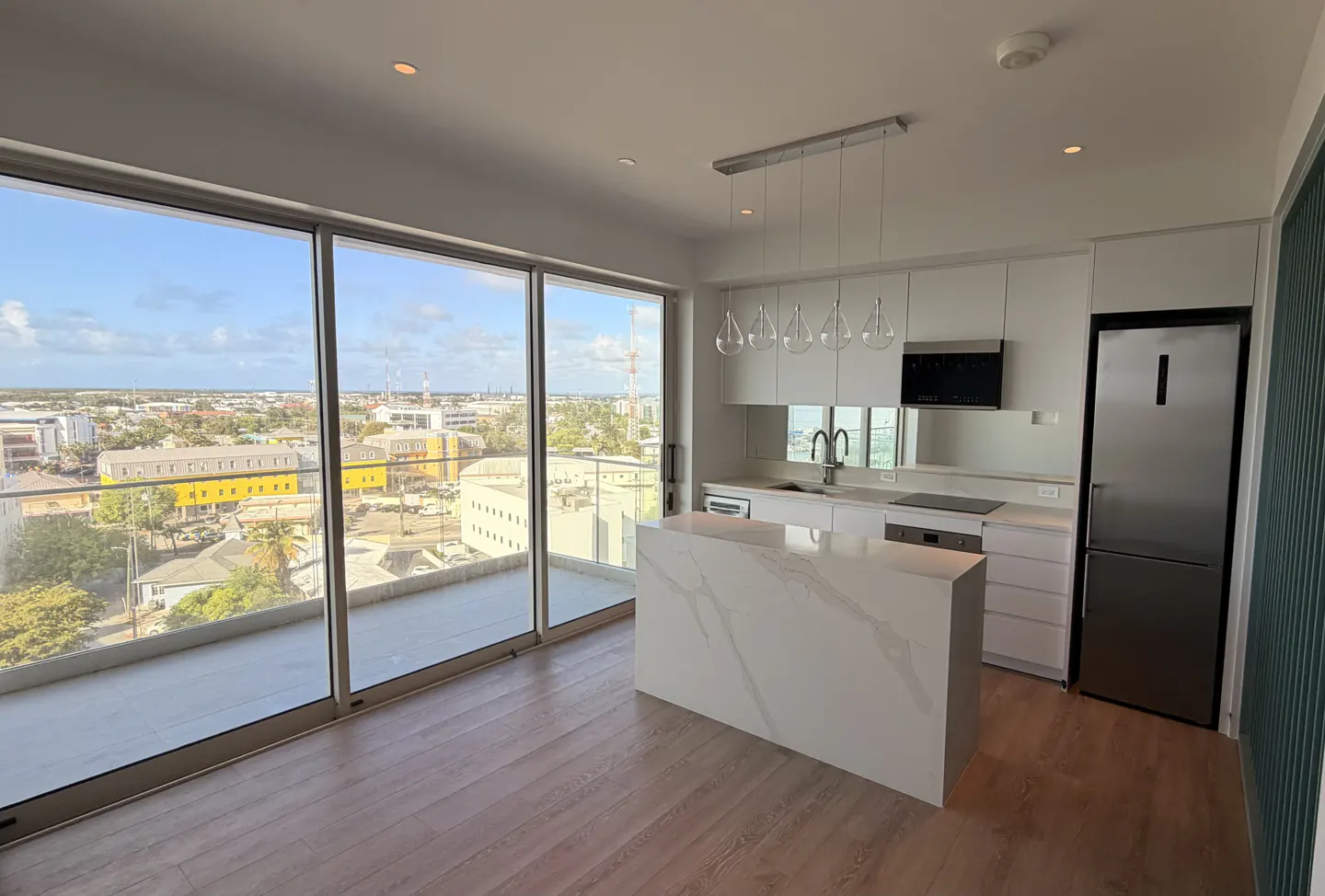 Bright, modern kitchen with white cabinets, marble island, and stainless steel fridge. Large windows offer a city view.