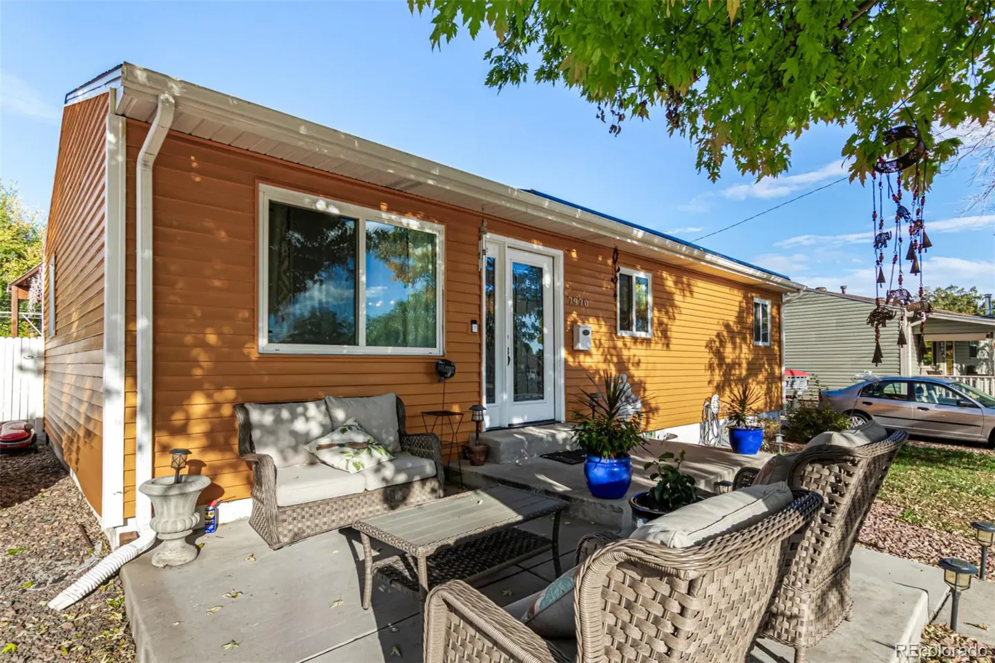 Exterior view of a one-story house with orange siding, white trim, and a patio with wicker furniture. Blue sky and green tree leaves are visible.
