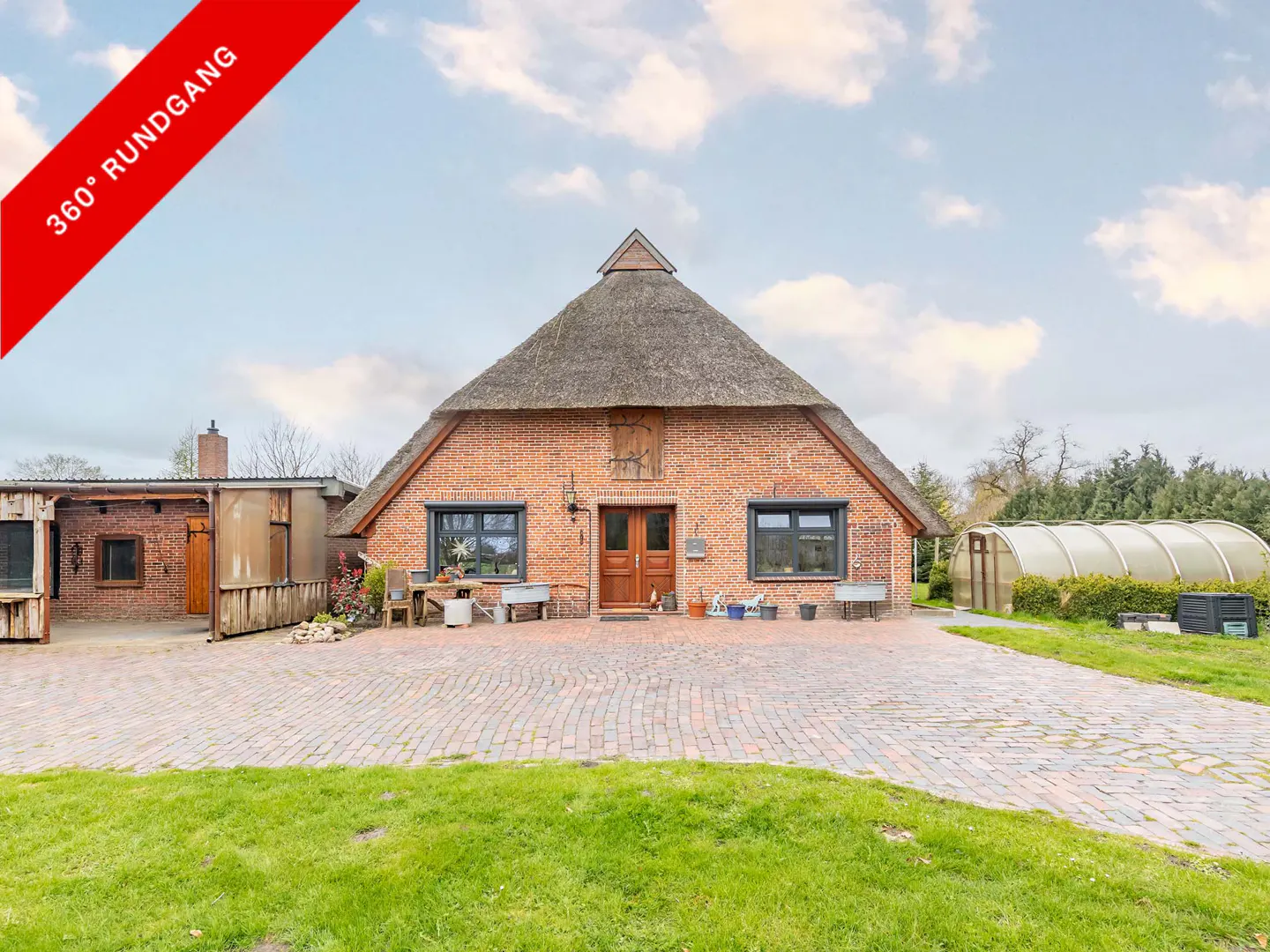 Exterior view of a red brick house with a thatched roof, a brick driveway, and a greenhouse in the background.