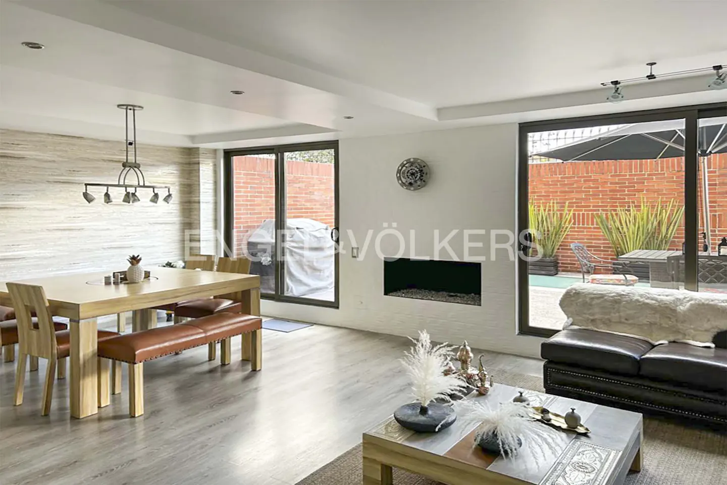 Modern open-plan living area with a wooden dining set, leather bench, fireplace, and sliding doors to a brick patio.