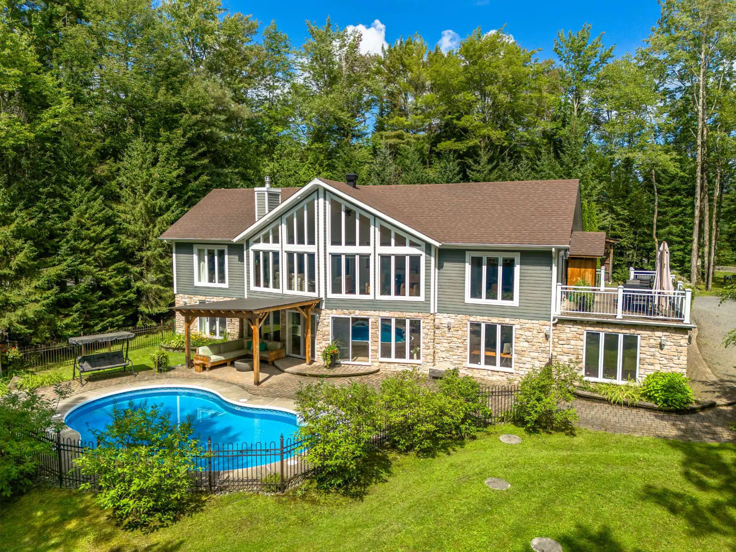 Two-story house with a pool. The house is gray with a brown roof and stone accents. The pool is surrounded by a black fence. Trees in the background.