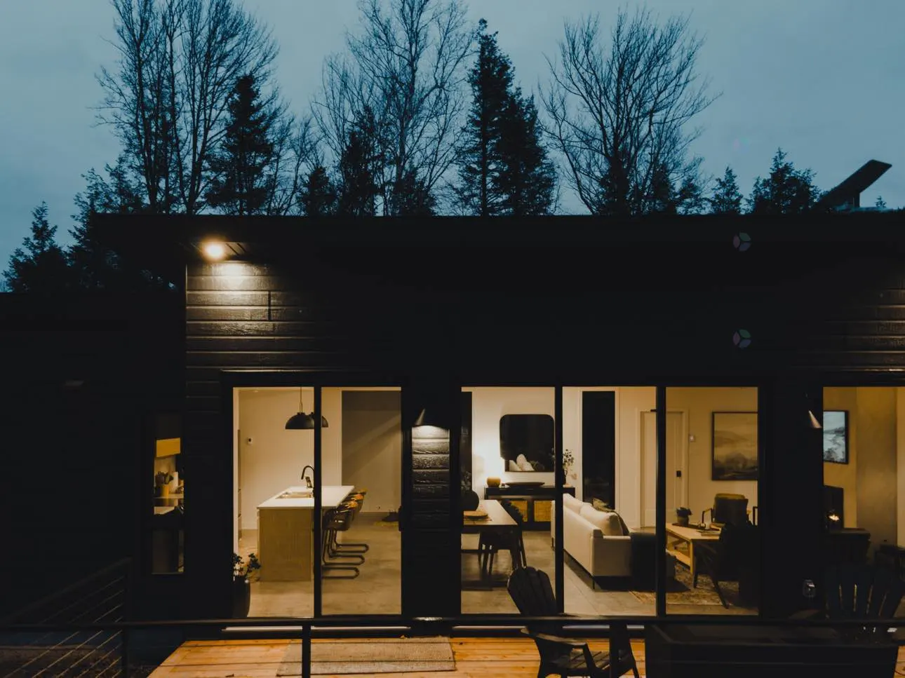 Exterior view of a modern, dark-colored house at dusk. Large windows reveal a bright, open-concept interior with a kitchen, dining area, and living room. Trees are visible in the background.