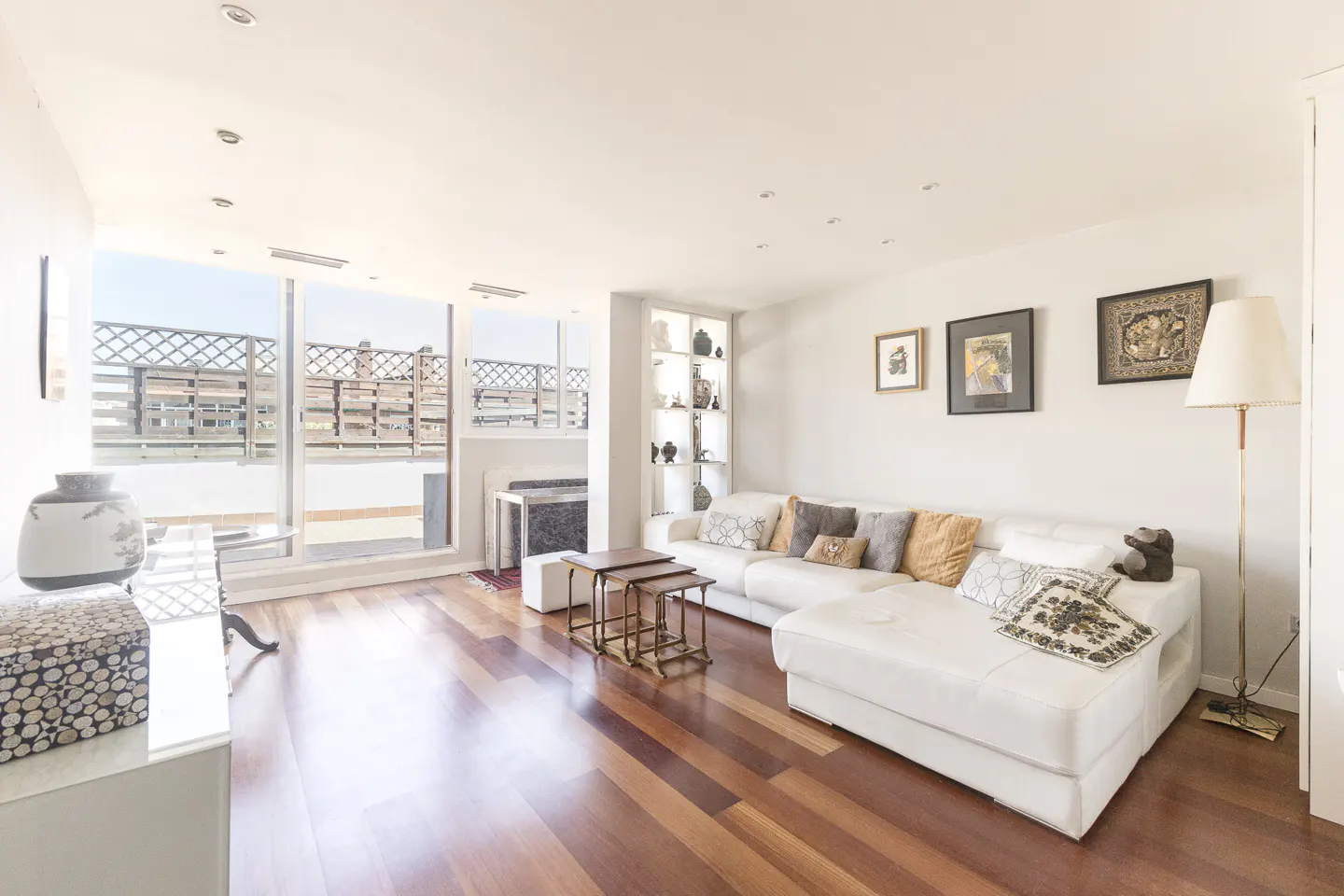Bright living room with hardwood floors, white walls, and a white sectional sofa with decorative pillows. Large windows overlook a balcony.
