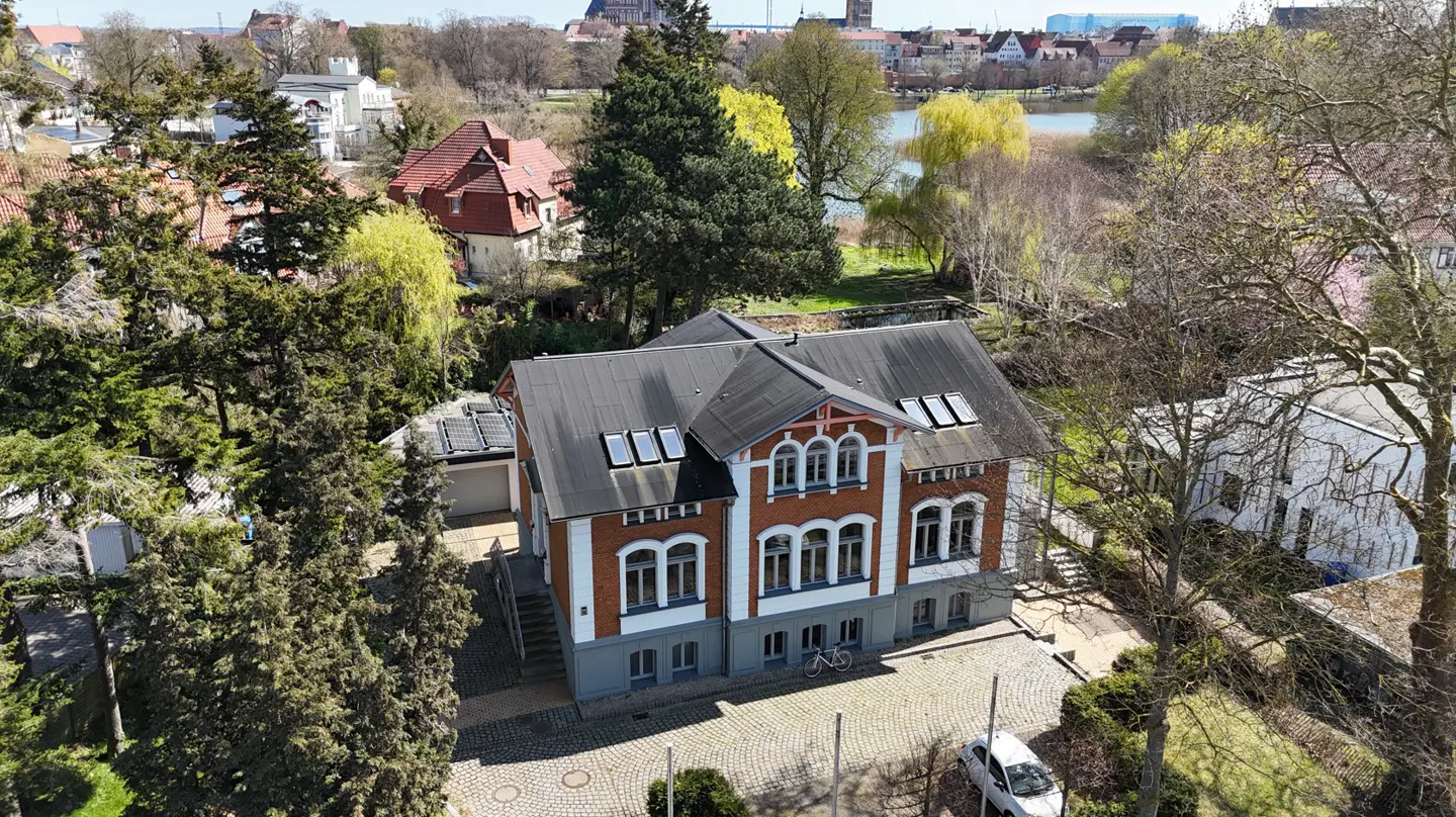Aerial view of a two-story brick house with a dark roof, white trim, and skylights, surrounded by trees and a town in the background.