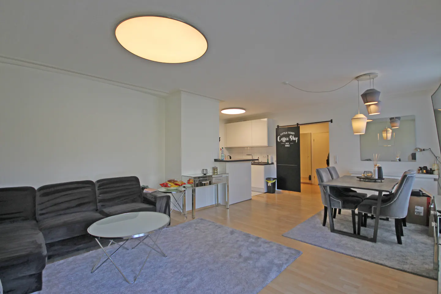 A bright, open-concept living and dining area with a gray sofa, dining table, and a kitchen in the background.