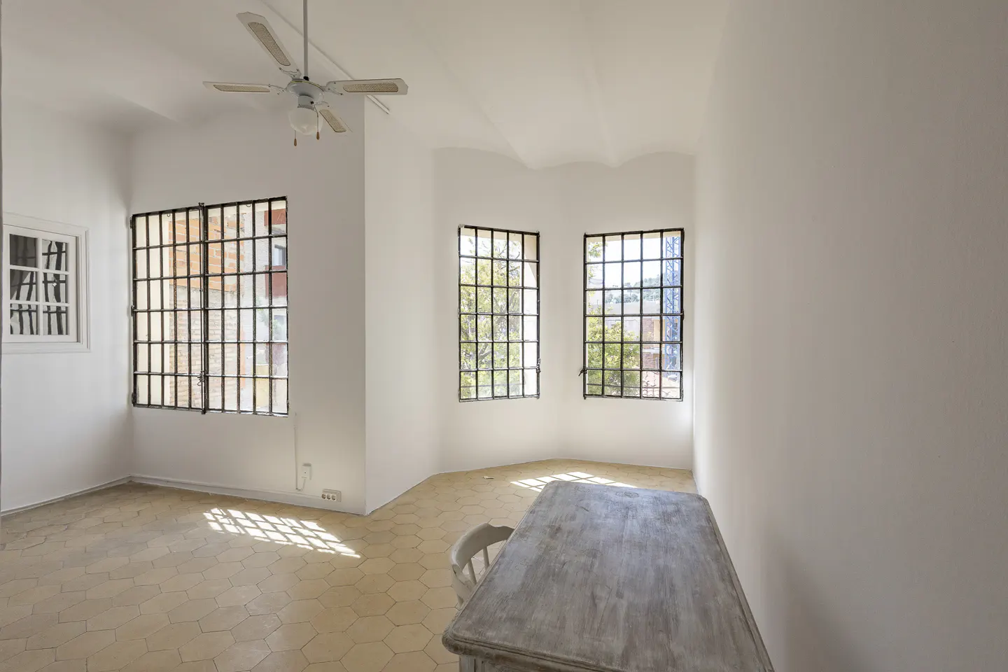 Bright, empty room with white walls, tile floor, and barred windows. A ceiling fan hangs above, and a distressed table sits in the foreground.