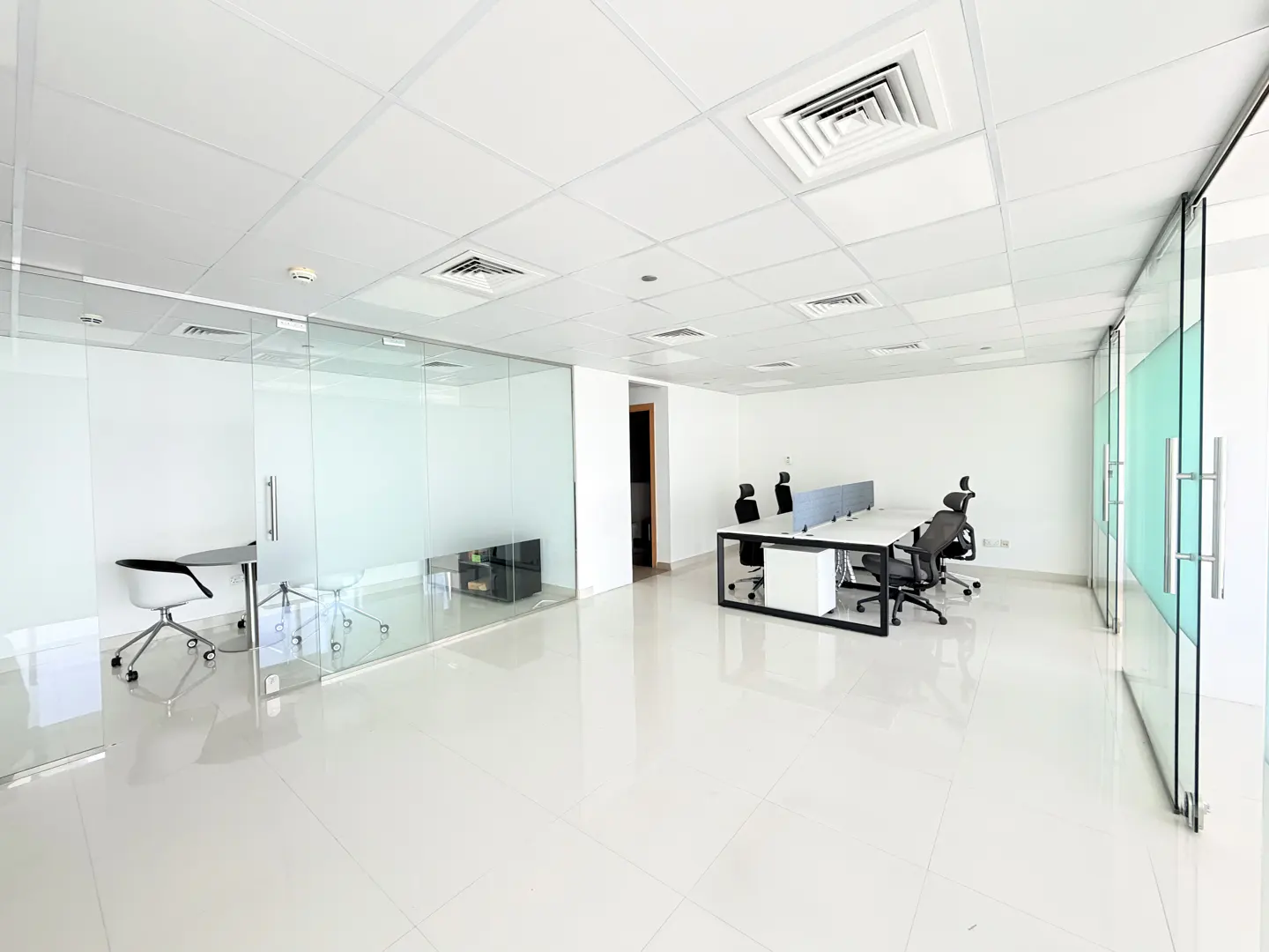 Bright, empty office space with white tile floors, glass walls, and modern desks with black chairs.