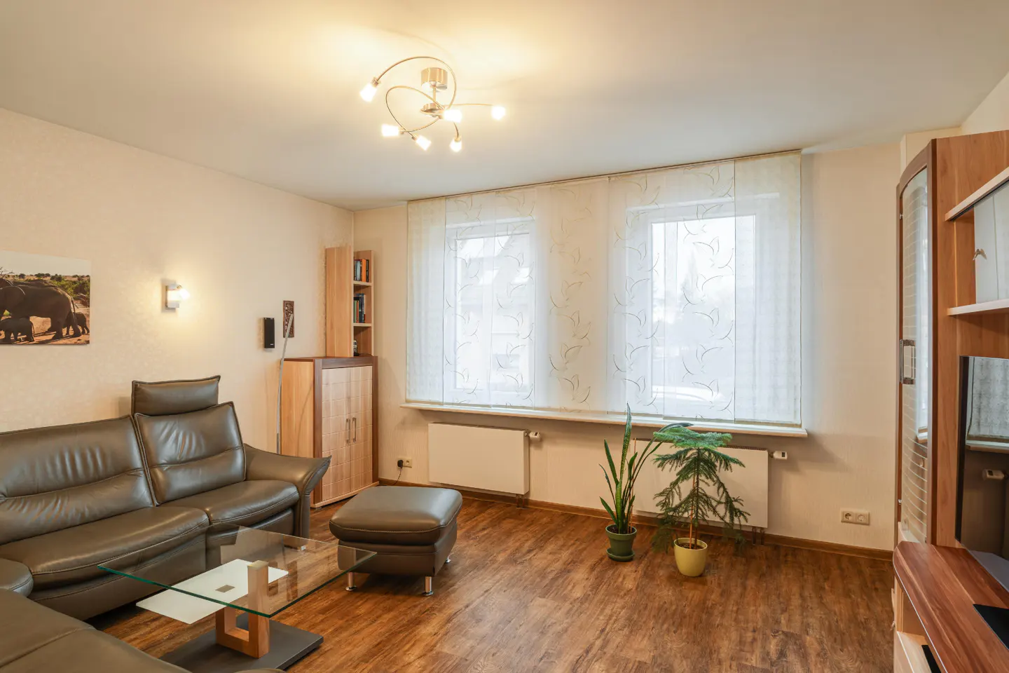 Living room with brown leather sofa, glass table, wood floors, and two windows with sheer curtains.