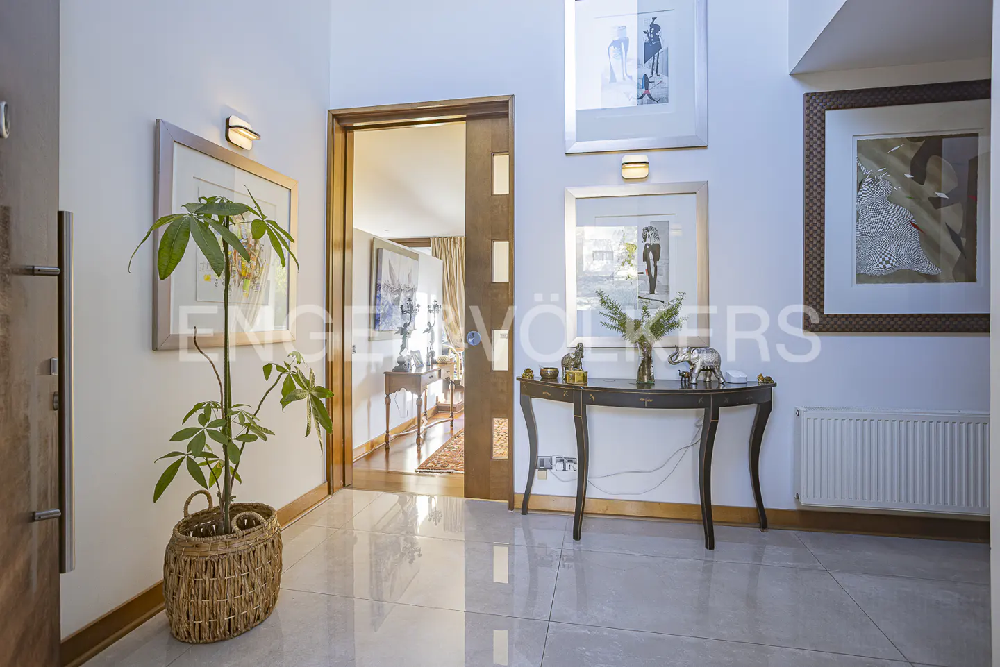 Foyer with gray tile floors, white walls, and artwork. A plant sits in a woven basket. A dark console table holds decorative items.
