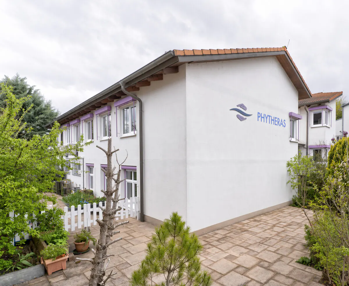 White two-story building with Phytheras logo, tiled roof, and a stone patio surrounded by green trees and a white fence.