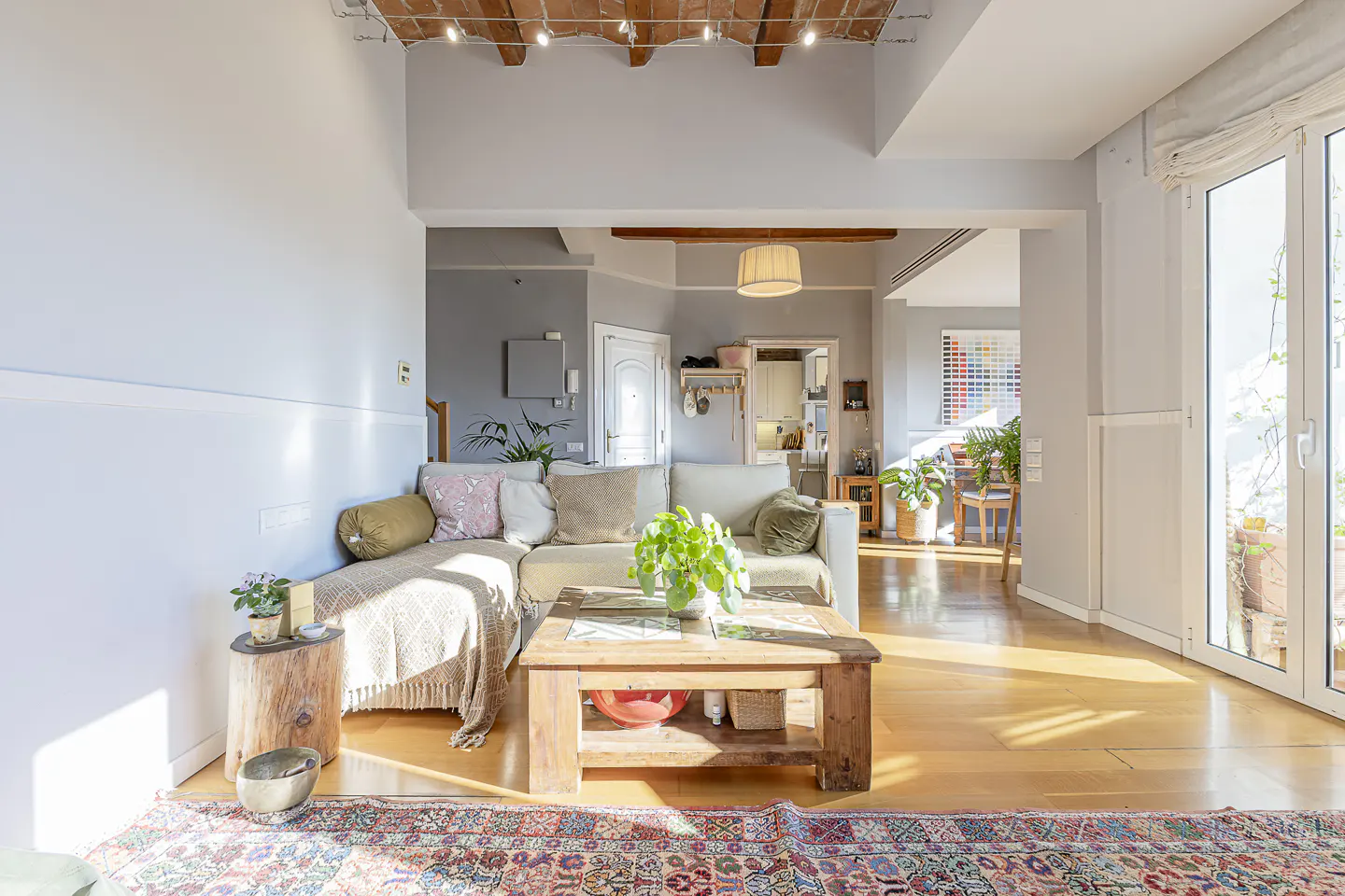 Bright living room with hardwood floors, a beige sectional sofa with pillows, and a rustic wooden coffee table with a plant. A colorful rug sits on the floor.