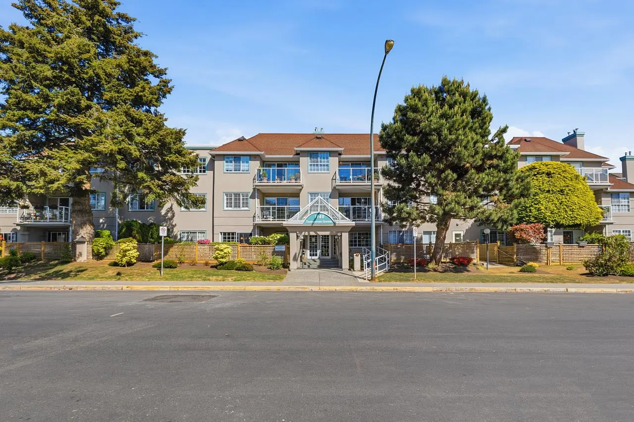 A three-story apartment building with balconies, a brown roof, and a glass entrance. Trees and a street are in the foreground.