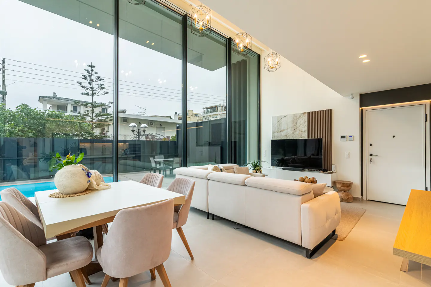 Bright living room with a white sofa, dining table with pink chairs, and floor-to-ceiling windows overlooking a pool.