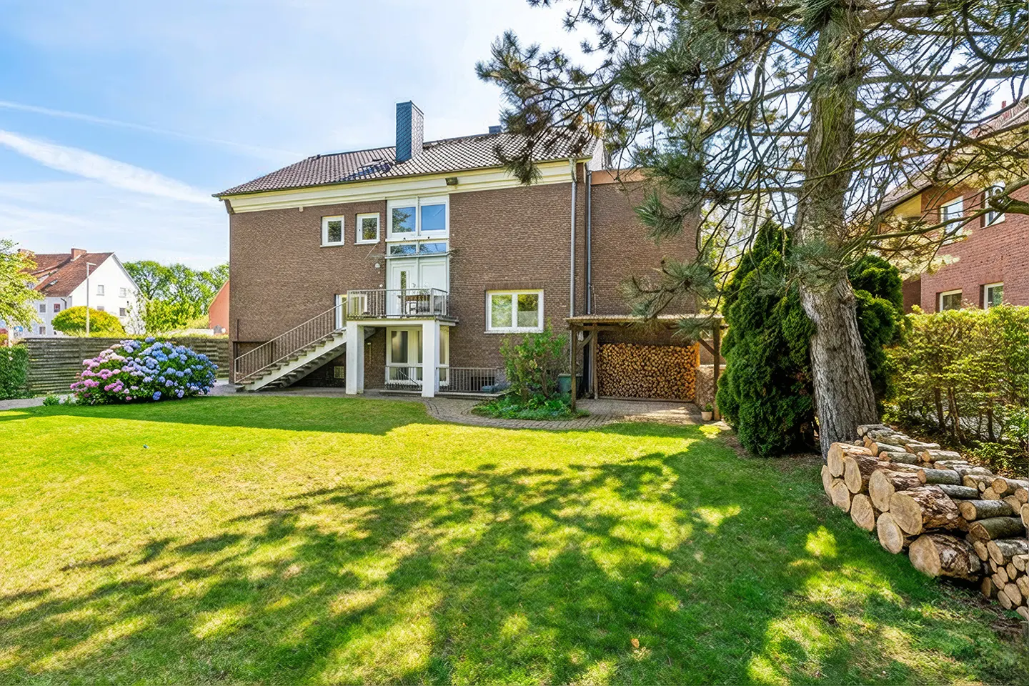 Backyard view of a two-story brick house with a balcony, stairs, and a wood storage shed. A green lawn and trees surround the house.