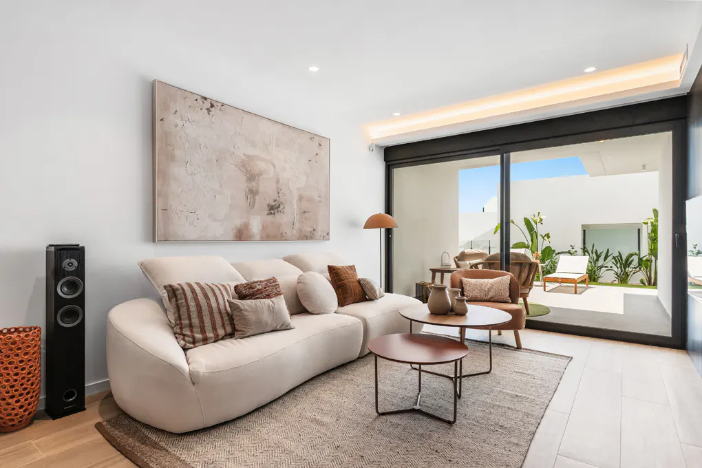 Living room with a white sofa, brown pillows, and a painting. A sliding glass door leads to a patio with plants and a lounge chair.