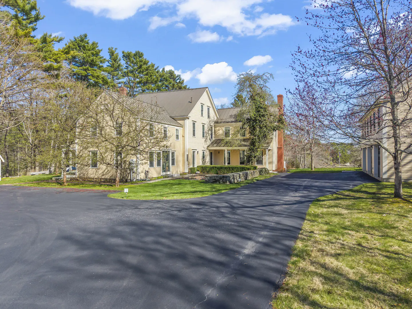 Exterior view of a large, light yellow house with a dark driveway and green lawn under a blue sky with scattered clouds.