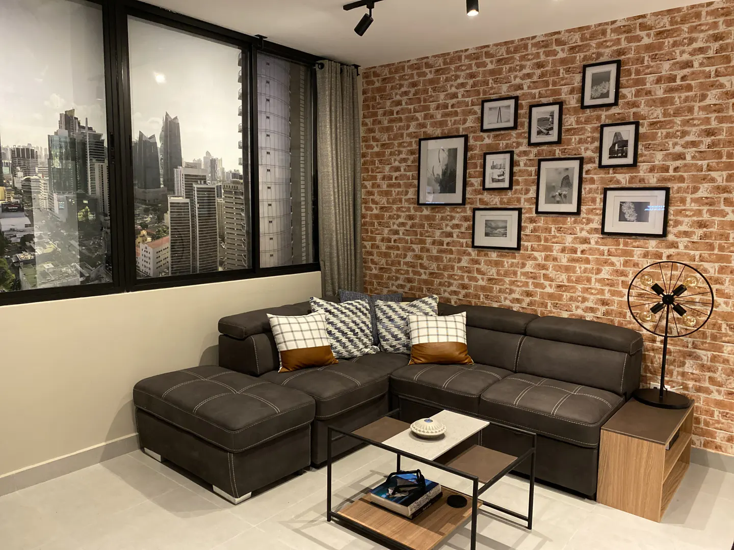 Modern living room with a grey sectional sofa, brick wall, gallery of framed photos, and a city skyline view.